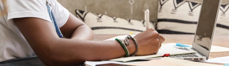 A person seated at a table, using a laptop while taking notes in a notebook.